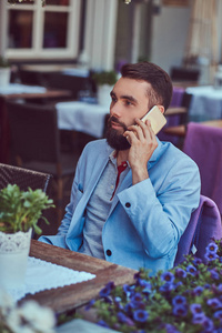 一个时尚的胡子男性的肖像, 时尚的发型, 电话交谈, 坐在户外咖啡馆