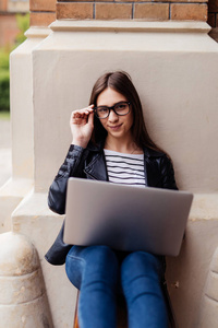 年轻的女性博客与追随者聊天, 并发送反馈休息在镇上的长凳上与上网本, 时髦的女孩工作自由职业者使用笔记本电脑享受户外花费时间