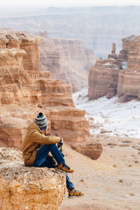 在哈萨克斯坦的 Charyn 峡谷, 一个带着照相机的欢快的旅行者坐在悬崖边上。