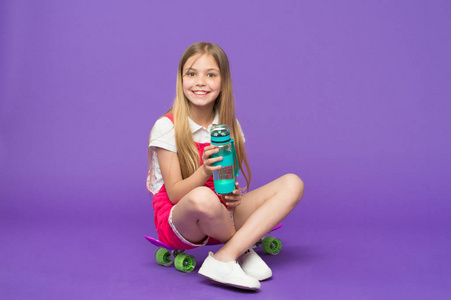 女孩在微笑的面孔举行与瓶水, 当坐在滑板, 紫罗兰色背景。长头发的小女孩喝水。水平衡概念。女孩关心健康和水平衡