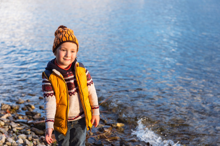 可爱的小男孩玩旁边湖在寒冷的日子，穿上暖和的套衫 黄色背心和帽子