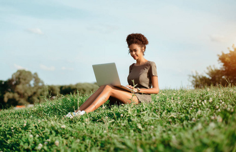 欢快的年轻非洲裔美国女学生与笔记本电脑在户外的草坪上的夏日巴西大学的女孩微笑着通过上网本做作业, 而坐在草地上