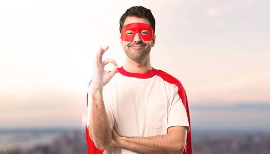 超级英雄男子与面具和红色斗篷显示一个 ok 标志与手指。在日落背景下面对幸福和满足