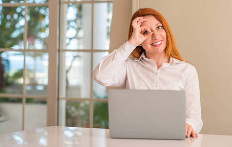 红头发的妇女使用电脑膝上型电脑在家里与快乐的脸微笑做 ok 签名用手在眼睛看通过手指