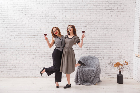 两个迷人的女孩，有乐趣和在聚会上喝红酒。时尚穿着漂亮的卷发。阁楼白色砖墙上
