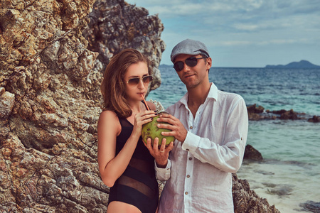有吸引力的情侣, 拿着一根管子的椰子, 站在靠近大礁石的海滩上, 在美丽的岛屿上享受假期。