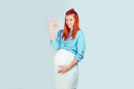 可爱的年轻孕妇穿着时髦的衣服轻轻抚摸腹部, 显示确定的手势看着在蓝色的相机隔离。我怀孕了, 一切都好。拉丁裔爱尔兰妇女