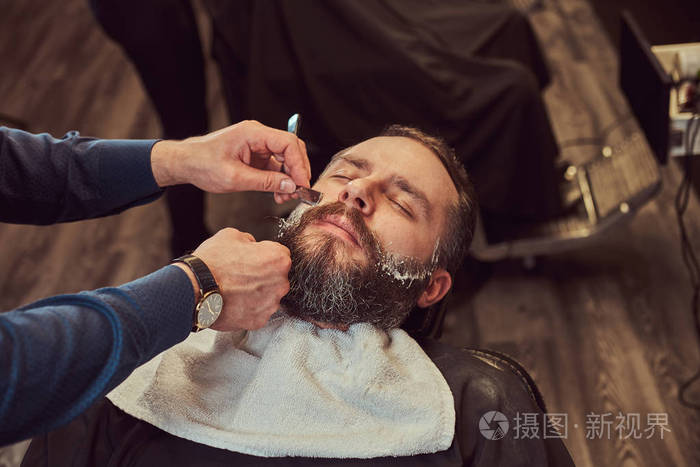 一个英俊的时髦胡子男子坐在理发店的扶手椅, 而理发师剃他的胡子与