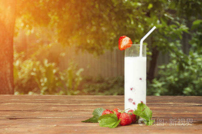 一杯带有新鲜牛奶饮料的杯子, 上面有草莓浆果和草莓叶子, 还有一张木桌和花园的背景。户外休闲的概念