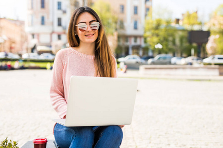 现代的博客。美丽的年轻女子在太阳镜使用她的笔记本电脑和视线笑而坐在户外阳光街的侧视图