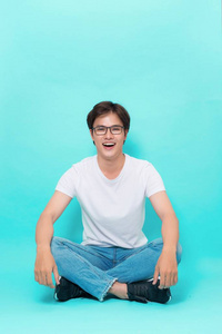 年轻的休闲亚洲男子坐在地板上, 他的腿交叉和微笑的相机。在蓝色背景上