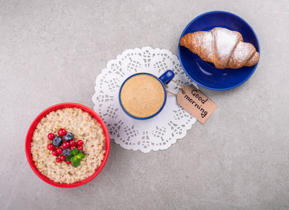 咖啡杯 浆果燕麦 羊角面包和注意早上好在灰色的背景上。健康早餐概念