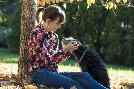 年轻女子坐在秋天的树下拥抱她黑色的狗