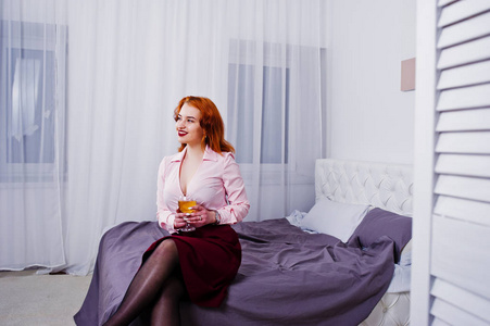 华丽的红头发的女孩在粉红色的上衣和红色的裙子, 手上拿着酒杯, 在房间的床上