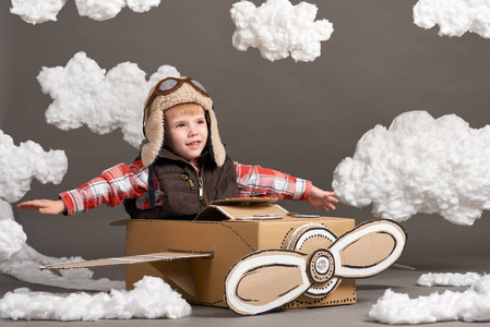 男孩在一架由纸板箱制成的飞机上玩耍, 梦想成为飞行员, 灰色背景下的棉绒云
