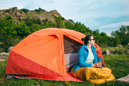 在森林中露营。那个女孩从附近山上背景帐篷热杯子喝咖啡。一个女人坐在附近一个帐篷睡袋中和从一个热水瓶喝茶