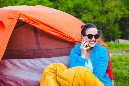 那个女孩用手机讲话。一个女人坐在帐篷旁边的一个睡袋里。远程通信。野营在自然。黑发手持智能手机