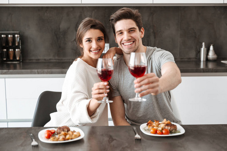 一对快乐的年轻夫妇的肖像, 坐在厨房的餐桌上, 并举行晚餐的酒杯