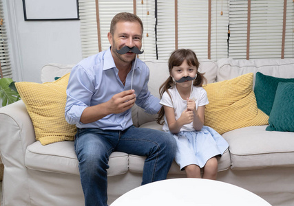 可爱的小女孩和她英俊的父亲与人造胡子在客厅看相机。父亲节快乐