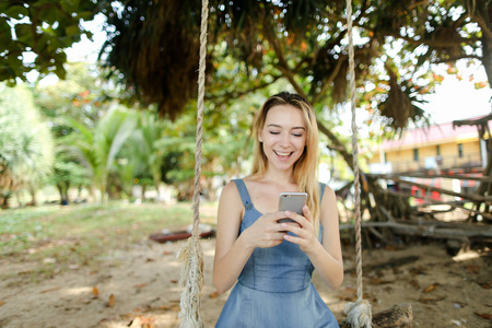年轻的微笑的女孩使用智能手机和享受社交网络, 骑在沙滩上摇摆