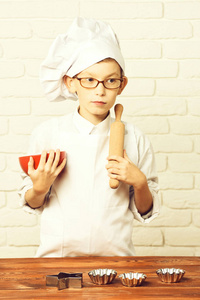 白色制服，戴着眼镜站附近表与模具蛋糕和持有用擀面杖上砖的背景墙上的大红碗副严肃的面孔上的帽子的小男孩小可爱厨师厨师