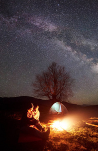 在山上露营的夜晚。一对徒步旅行的男人和女人在星空下, 银河中的燃烧篝火旁休息。照明帐篷, 大树和远处的丘陵背景。户外活动概念