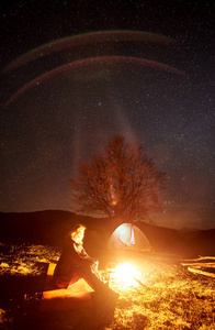 在山上露营的夜晚。在黑暗的星空下, 坐着年轻的女性徒步旅行者, 在风景灿烂的天空下, 带着旅游帐篷和远处的山丘。旅游与户外活动概