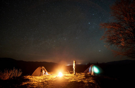 晚上露营在山上。年轻的旅游妇女站在两个照明帐篷之间的燃烧篝火附近的大树, 看着天空与许多明亮的闪闪发光的明星。旅游和旅游概念