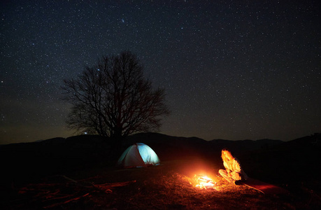 在山上露营的夜晚。在美丽的星空下, 年轻的女性游客在燃烧的篝火旁休息, 背景是明亮的帐篷和大树的剪影。户外活动概念