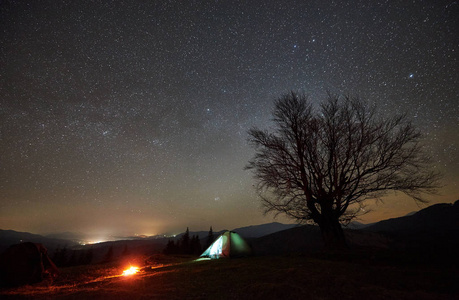 篝火附近的旅游照明帐篷燃烧, 从里面点燃。夜间露营在山下惊人的星空。远处的山丘, 背景大树的剪影。旅游业和旅游概念