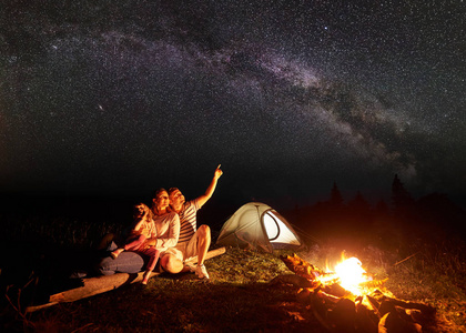 一家人晚上在山上露营, 坐在灯火通明的帐篷前, 燃烧着篝火。女子抱在怀里的女孩, 男子指着明亮的星星在黑暗的天空。旅游业和旅游概