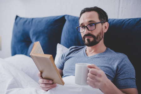 戴眼镜的男人在床上看书和捧着一杯茶