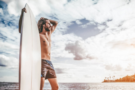 留着长头发的年轻英俊的男人正站在海滩与白色冲浪板在手上