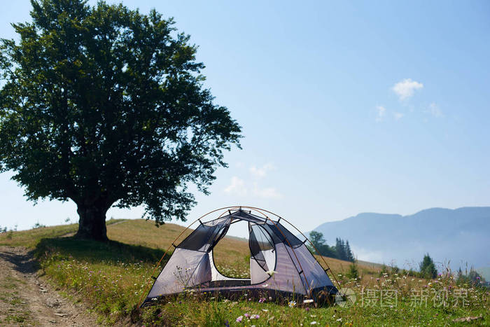 夏日美景的小舒适现代白色旅游帐篷在绿草山下的蓝天下, 大树的背景。野营旅游和旅游概念