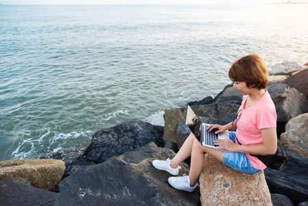 年轻的亚洲妇女自由职业者穿着粉红色衬衫坐在岩石上, 并与笔记本电脑在户外工作。海日落背景