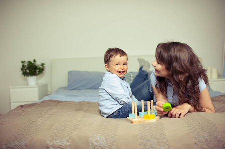 年轻的母亲和年幼的儿子坐在床上。妈妈和她的儿子一起玩。家庭