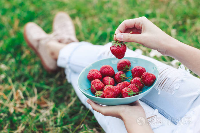 坐在夏草及持有一盘草莓 膝盖和手可见的牛仔裤的女孩。健康的早餐，干净吃纯素食品的概念。顶视图。色调