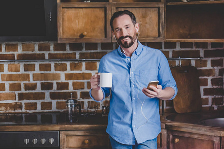 微笑中年男子拿着咖啡杯和听音乐在智能手机在厨房