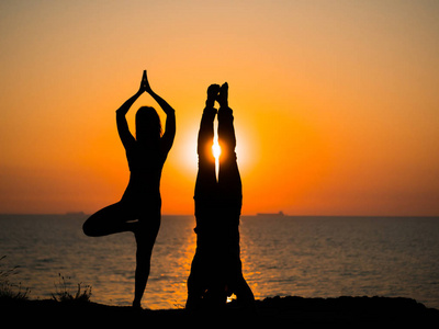 在日落的海边海滩上, 年轻女子和男子做双瑜珈的剪影。冥想。夫妇练习瑜珈。在自然背景下的柔韧性锻炼