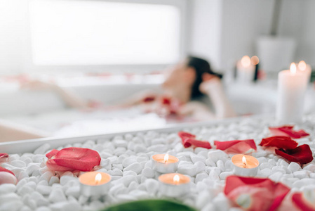 年轻女子躺在浴缸与泡沫, 玫瑰花瓣和燃烧蜡烛装饰照片