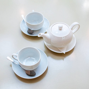 两个白色的陶瓷茶杯, 上面有闪亮的勺子和碟子, 咖啡桌上有茶壶。