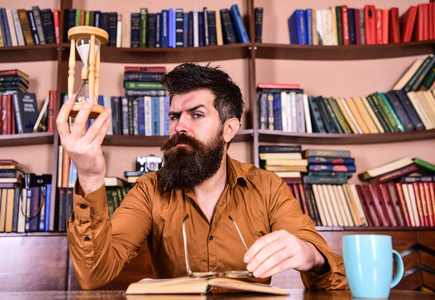 男人在忙碌的脸上看书, 书架上的背景。课和时间概念。在图书馆学习胡子的老师或学生。戴眼镜的科学家坐在桌子旁, 看着沙漏