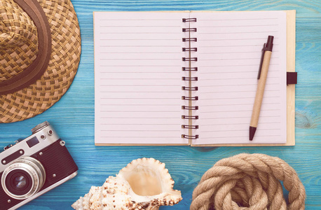 夏天的背景。夏天做的清单。空白页笔记垫贝壳绳索帽子和老式相片照相机在蓝色木桌上
