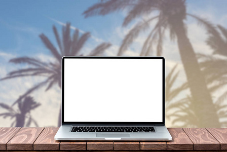 现代笔记本电脑与空白白色屏幕上的木桌反对模糊的棕榈树背景