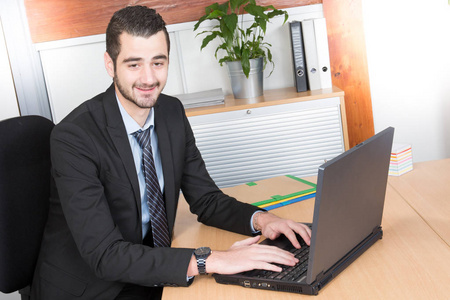 一个漂亮的微笑的商业人的肖像在办公室坐在办公桌上看着他的笔记本电脑