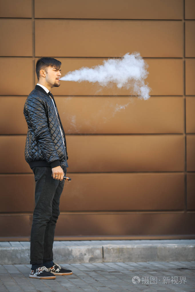 年轻白人帅哥与现代的发型 vaping 对面的未来派的城市背景的一种电子
