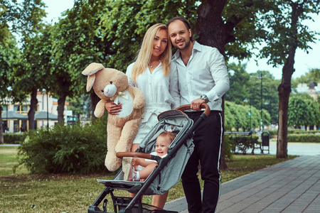 幸福的年轻家庭概念。美丽的金发母亲和胡子时尚的父亲走在公园与他们的小女儿在婴儿推车