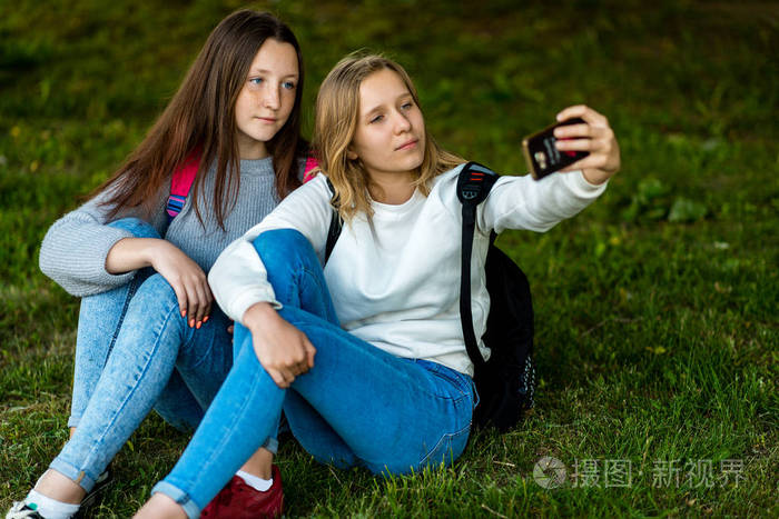 两个女朋友女生.夏天在公园在自然.坐在草地上, 背包里.手持智能手机.