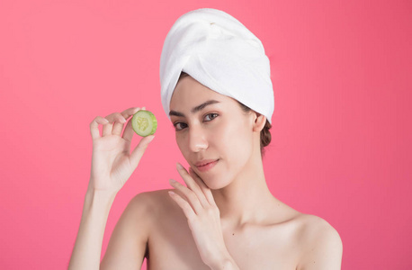 美丽的亚洲妇女与毛巾在头部与完美的清洁皮肤微笑着举行黄瓜片粉红色背景。美容美容水疗中心