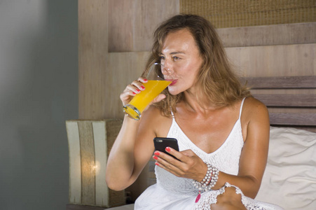 年轻迷人和美丽的女人在家里在床上使用互联网社交媒体应用程序在手机上微笑快乐饮用健康橙汁放松